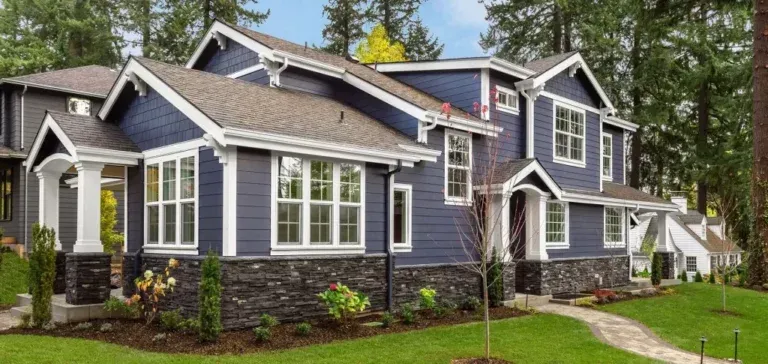 Blue-gray suburban house with white trim, gabled roof, and landscaped yard beside tall trees