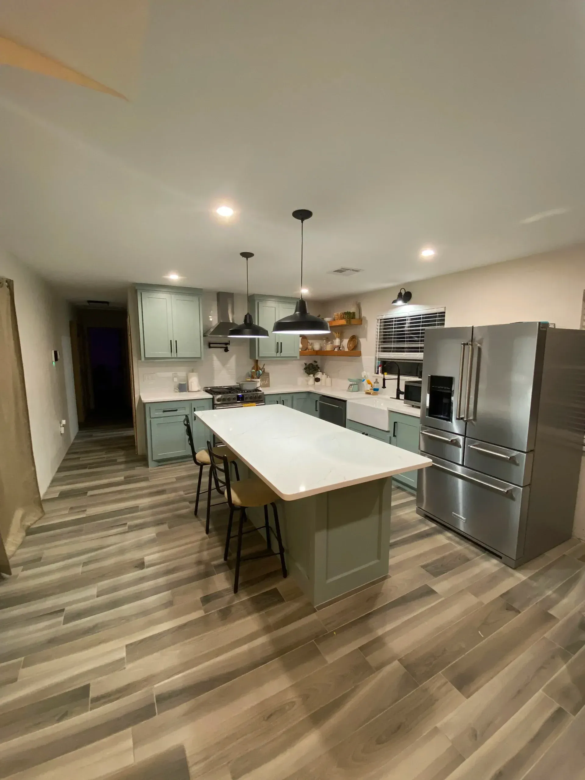 Modern kitchen with large island, stainless steel refrigerator, wood floors, and pendant lighting.