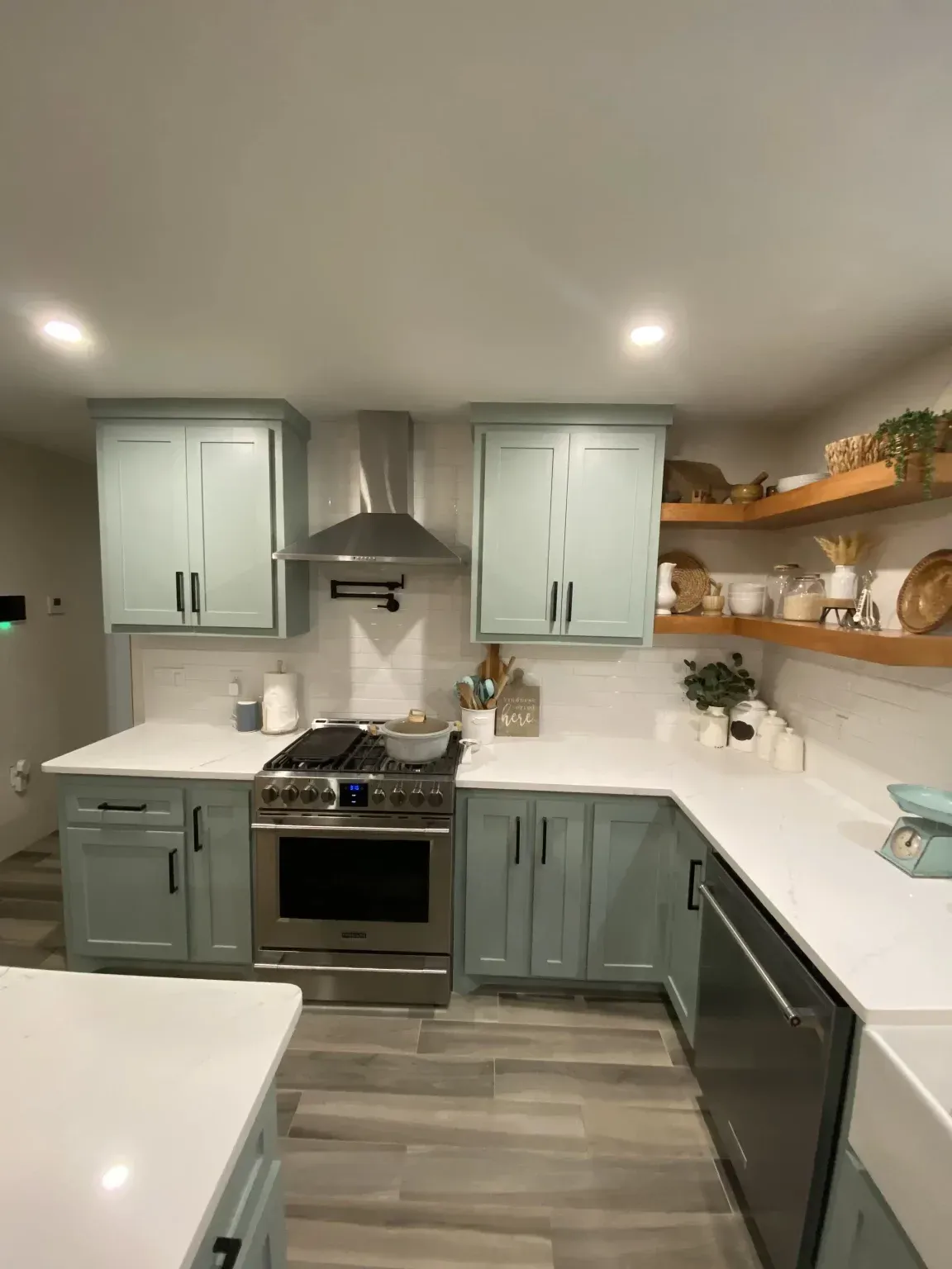 Modern kitchen with sage cabinets, white countertops, stainless appliances, and open shelves with decor.