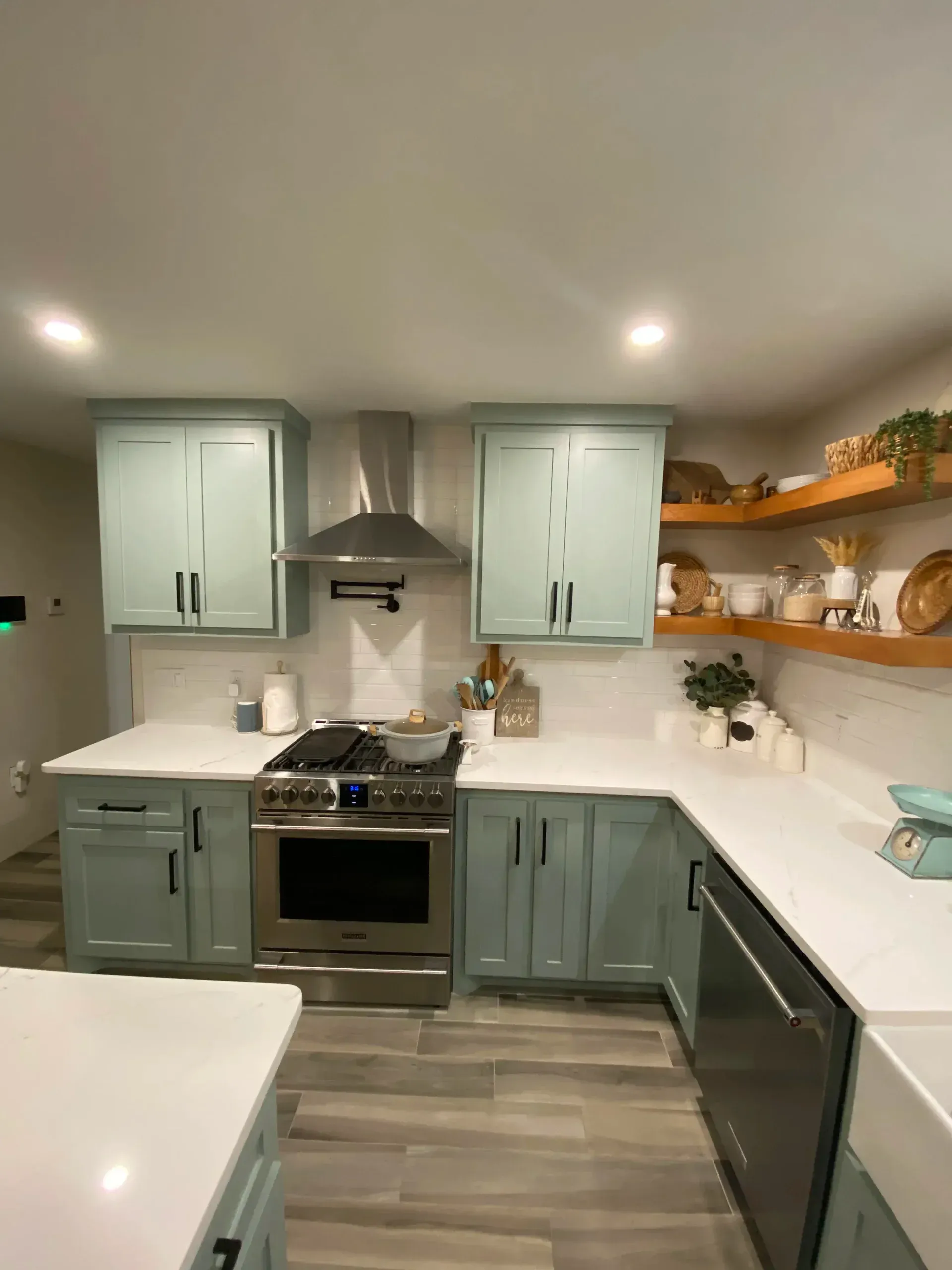 Modern kitchen with sage green cabinets, white countertops, stainless stove, and open wooden shelves