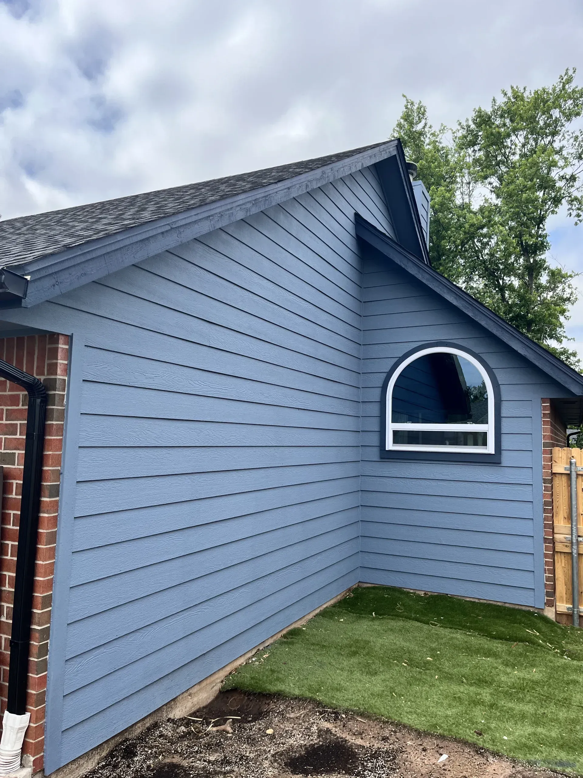 Blue-gray house side wall with an arched window, brick corner, and small grass yard under a cloudy sky
