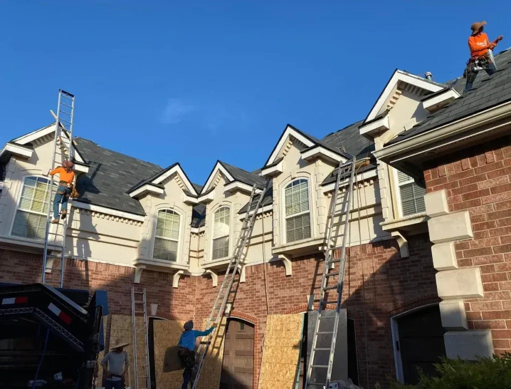 Workers on ladders repairing a brick townhouse roof under a clear blue sky.