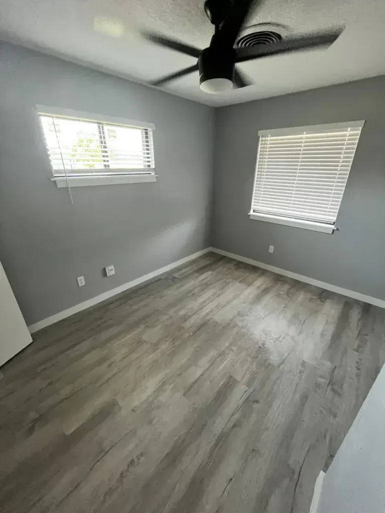 Empty gray bedroom with wood-look flooring, two windows with blinds, and a ceiling fan.