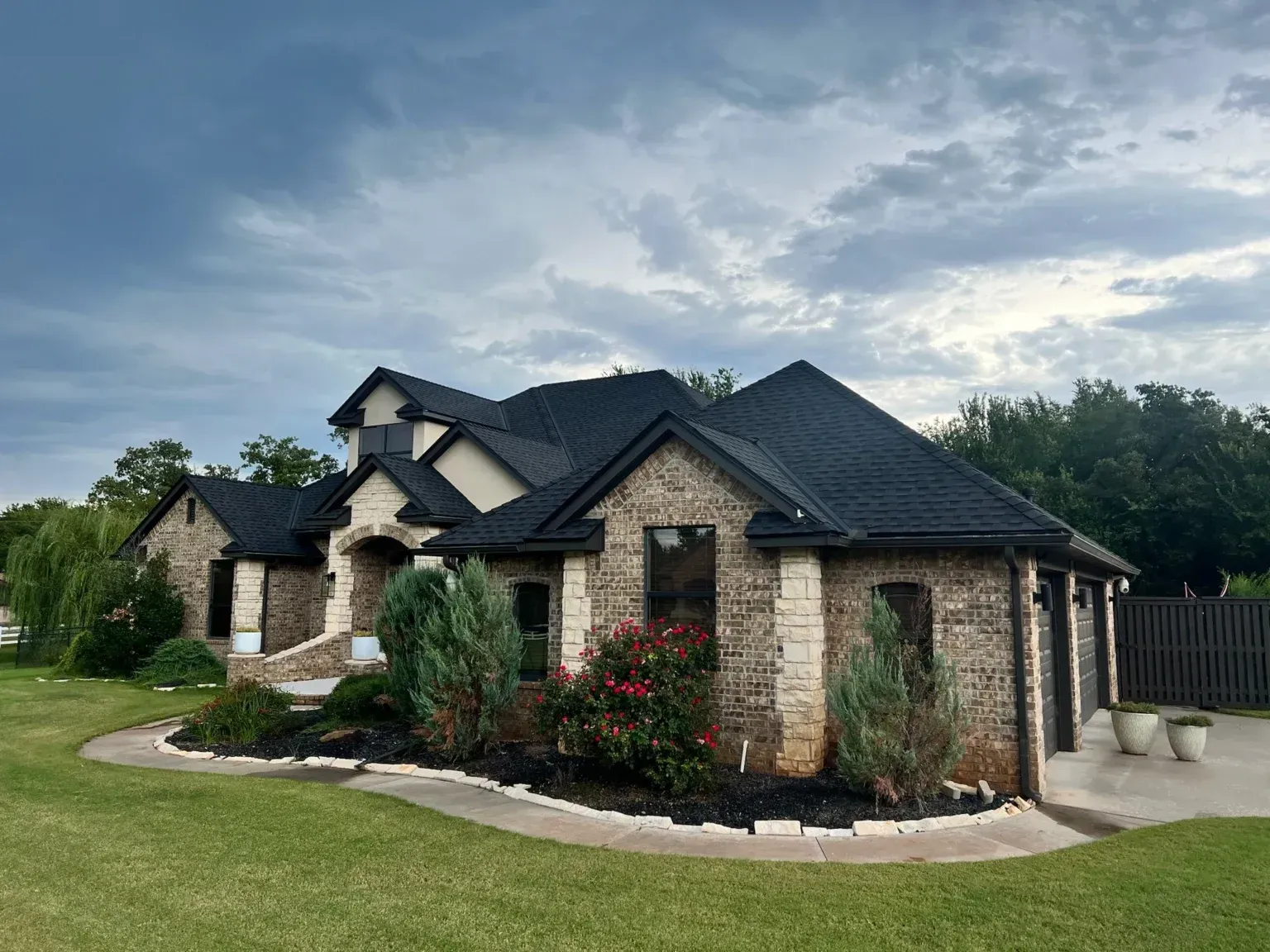 Single-story stone house with dark roof, landscaped yard, and cloudy sky
