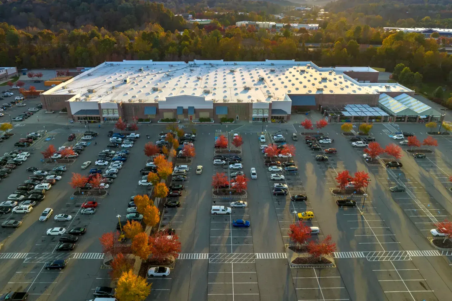 Aerial view of a large parking lot beside a big white-roofed store with autumn trees around it