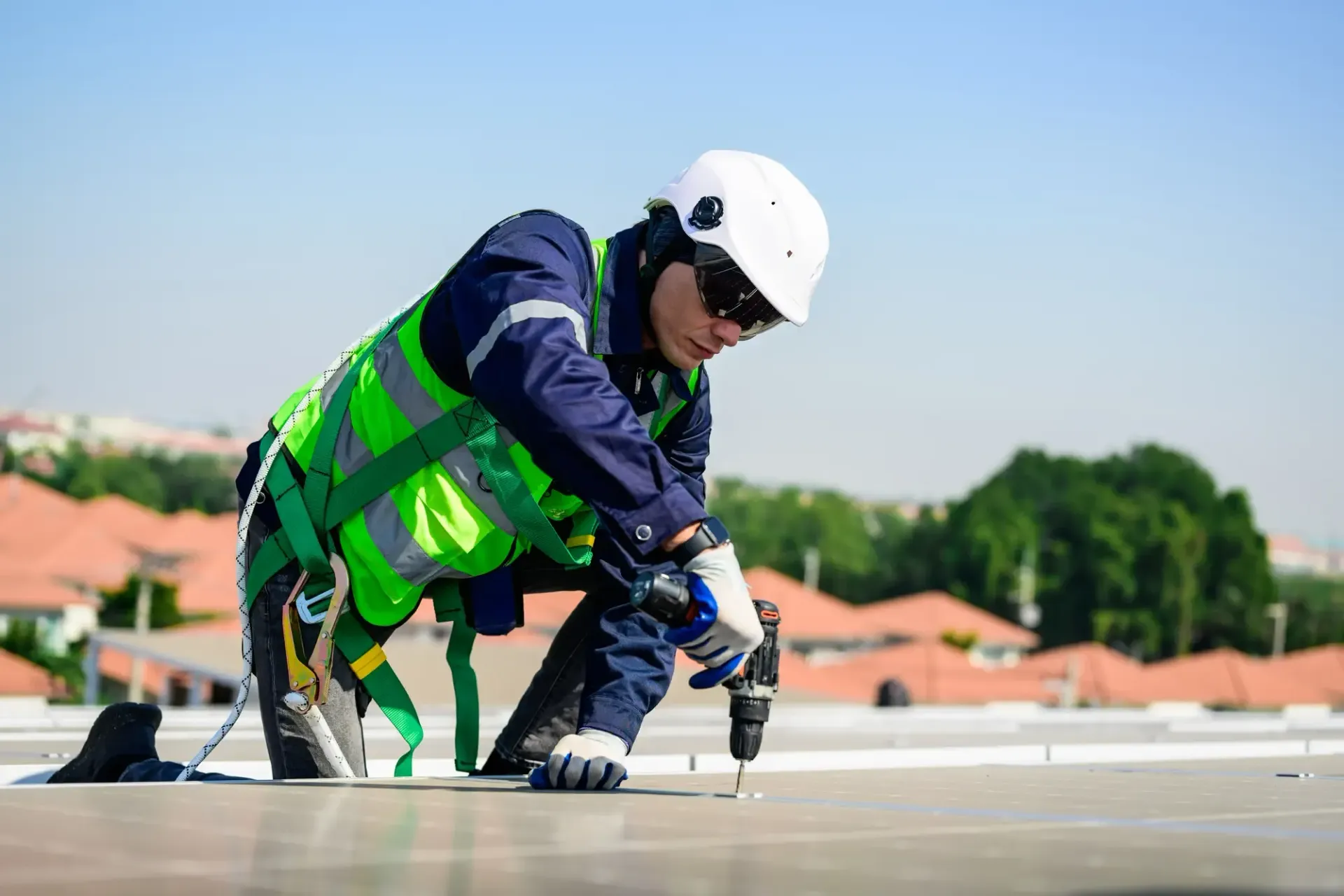 Construction worker in green safety vest and white helmet kneeling on a rooftop, using a power tool.