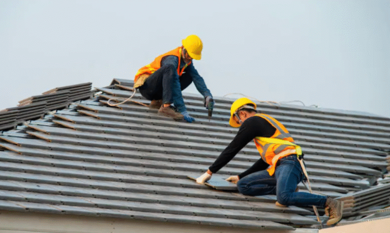 Two roof workers in orange vests and yellow hard hats repairing a gray shingled roof