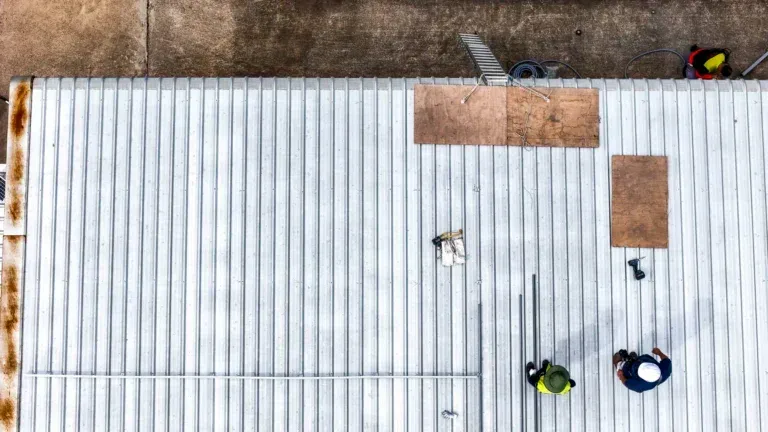 Workers on a white metal roof patching damaged sections, viewed from above.