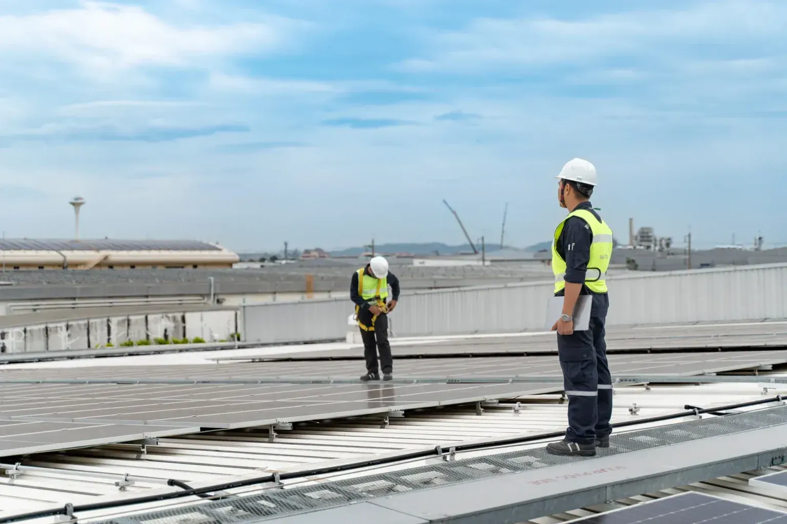 Construction workers in hard hats inspect rooftop solar panels under a partly cloudy sky.