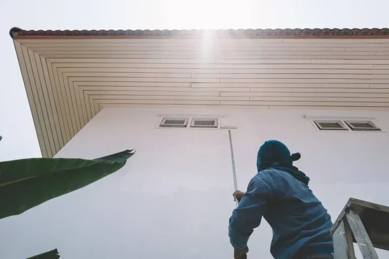 Person in blue hoodie holding a long pole, reaching up to a white house wall under the eaves.