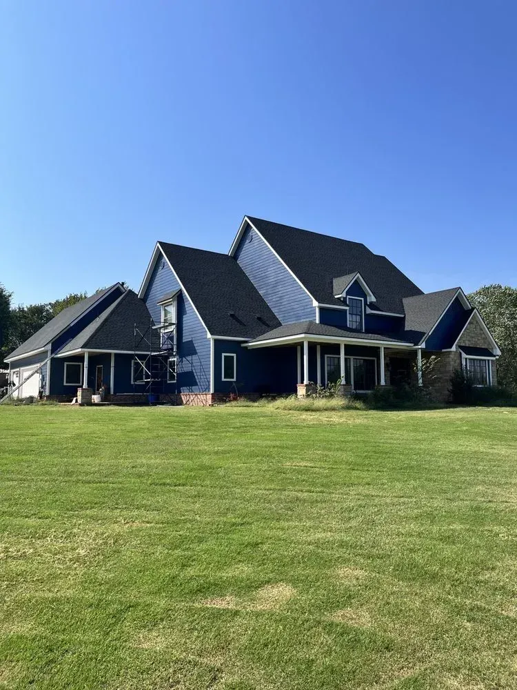 Large dark-blue house with gabled roofs and a covered porch on a wide green lawn under a clear blue sky