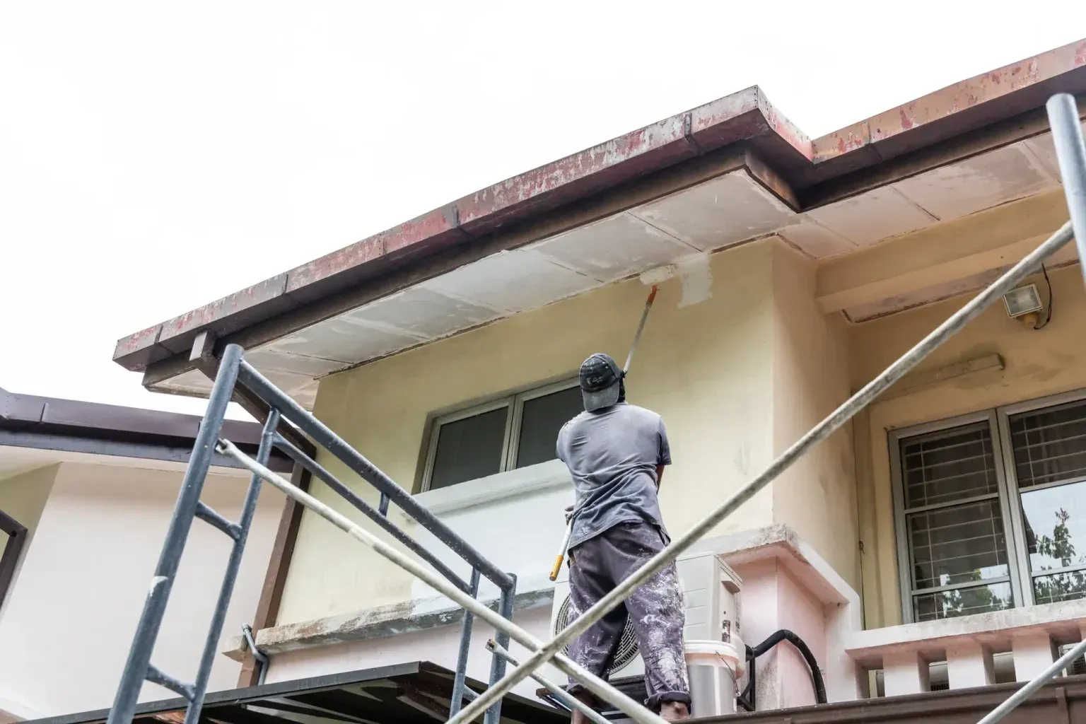 Worker painting exterior wall from scaffolding under the roof eaves