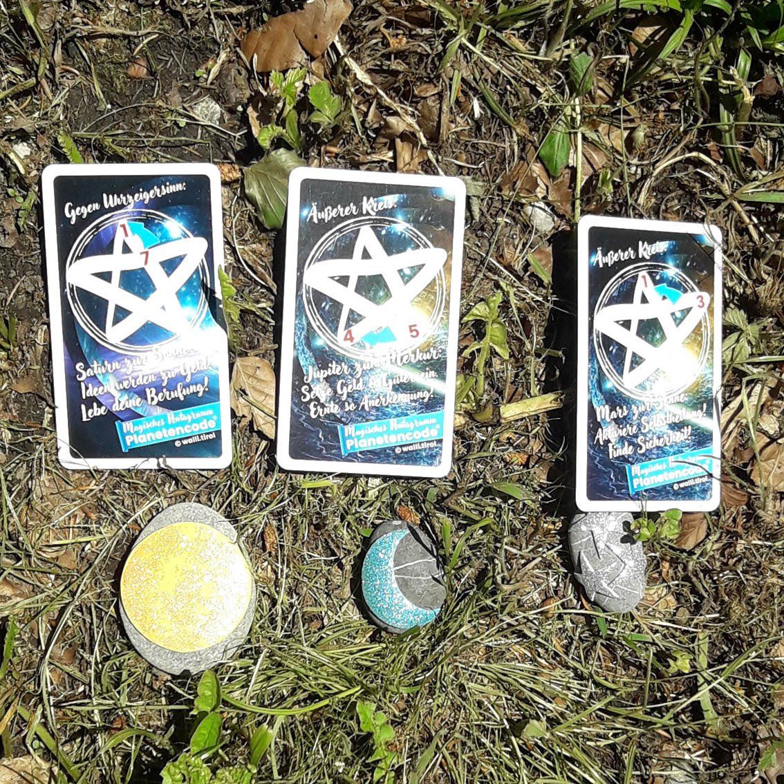 Drei tarotähnliche Karten und drei kleine Steine ​​auf Gras und Erde im Freien.