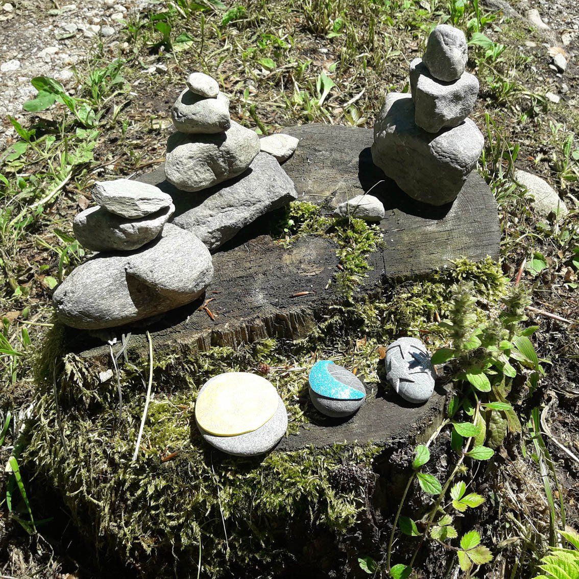 Kleine Steinhaufen auf einem moosbewachsenen Felsen im hellen Sonnenlicht, davor drei bemalte Kieselsteine.