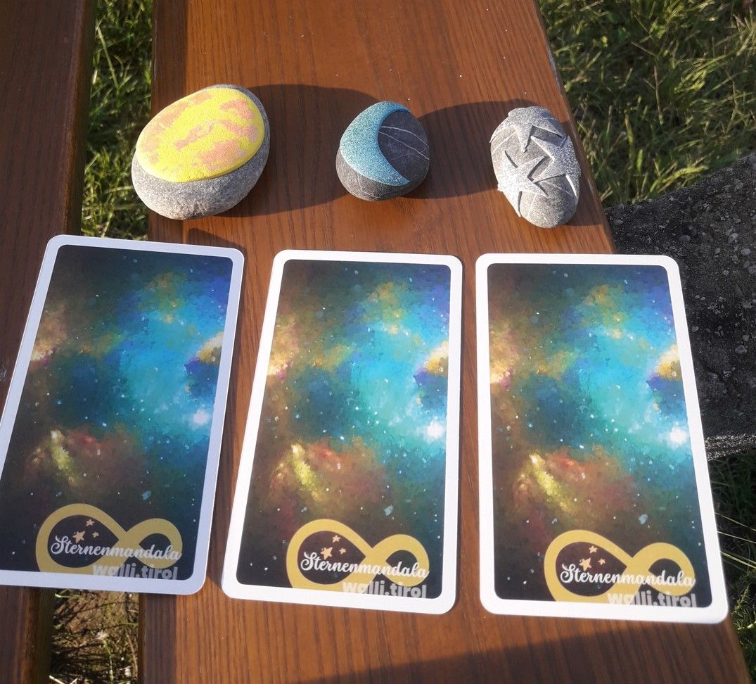 Drei Tarotkarten lagen im Sonnenlicht auf einem Holztisch neben drei bunten Steinen.