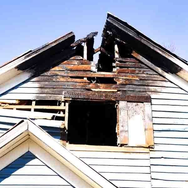 Damaged house gable with a gaping hole and charred wood against a blue sky.
