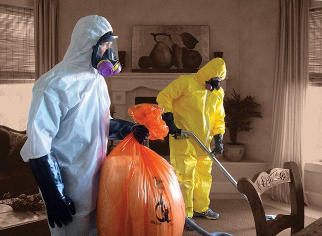 Two people in hazmat suits cleaning a living room; one holds a biohazard bag, the other vacuums.