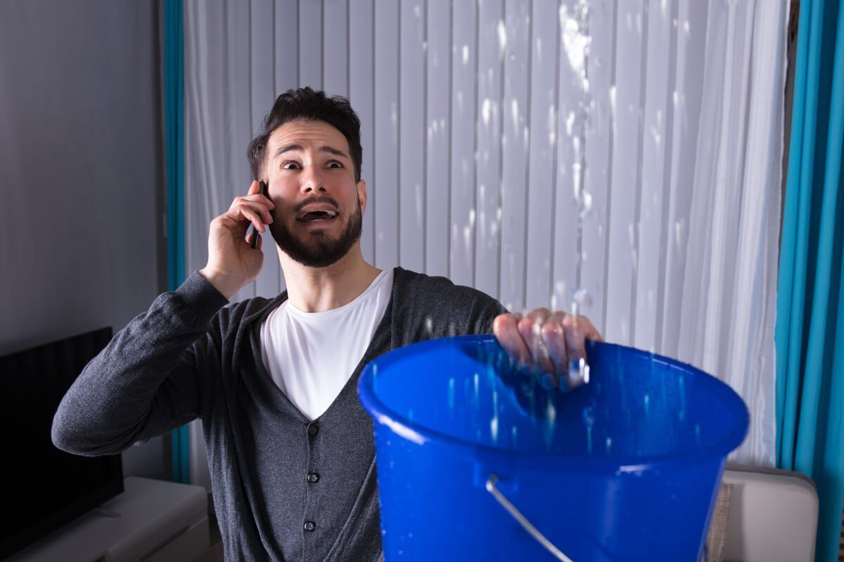 Man on phone, holding bucket to catch water leaking from ceiling. Interior scene.
