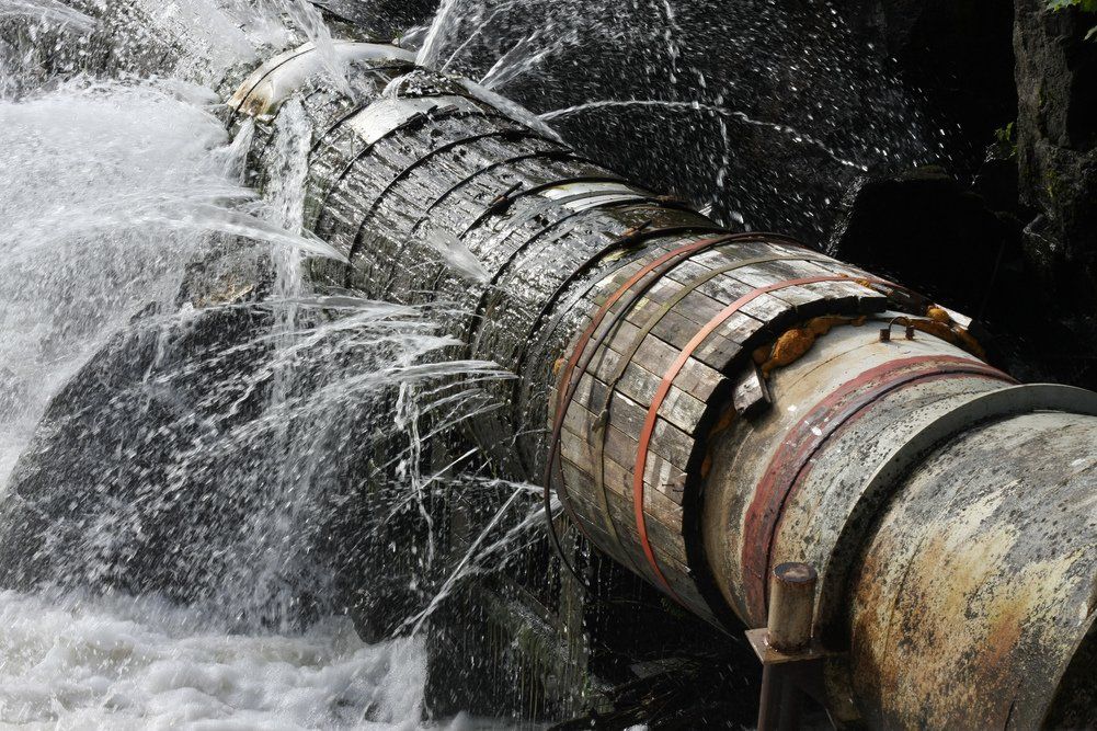 Water gushing from a damaged, rusty pipe in a rocky waterfall setting.