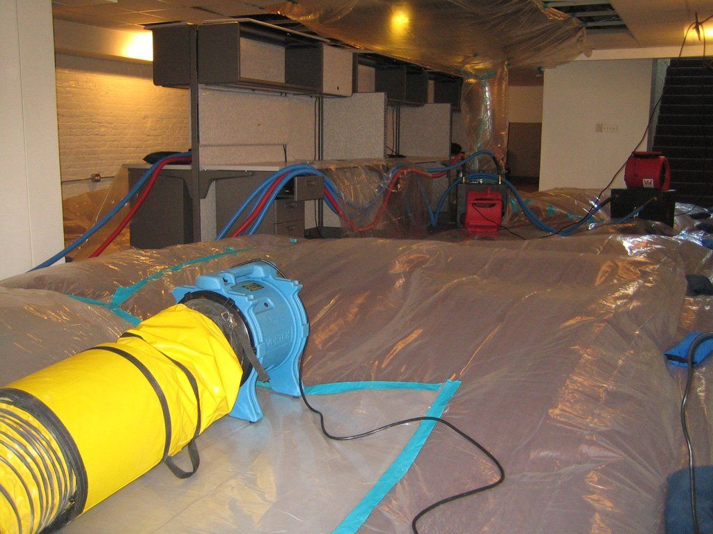 Interior view of a water damage remediation site. A large plastic containment area surrounds damaged furnishings. A blue fan and tubing are in use.