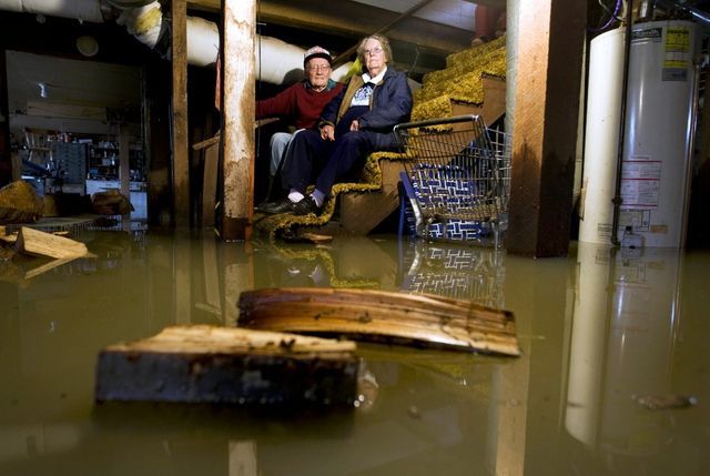 Two people sit on stairs in a flooded basement. Water and debris surround them.