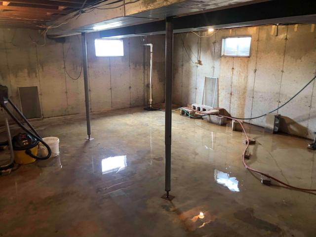 Flooded basement with concrete floor, metal support beams, and small windows. Water reflects light.