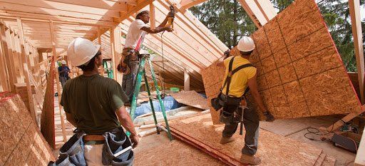 Construction workers installing plywood on a roof frame. One worker on a ladder uses a nail gun.