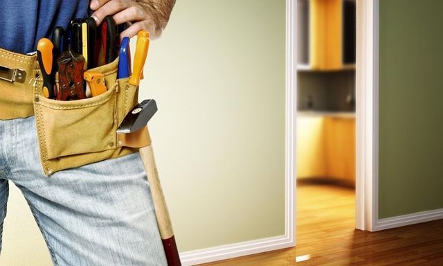 Man in jeans with tool belt, holding a hammer, doorway in background.