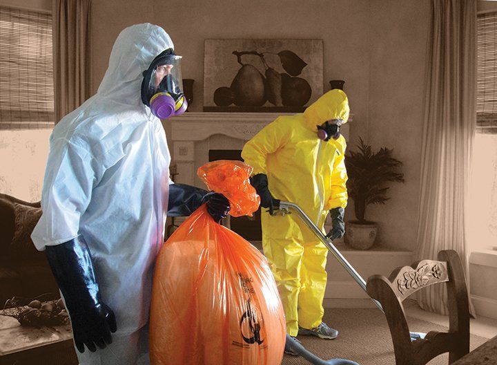 Two people in hazmat suits cleaning a living room; one holds an orange biohazard bag, the other uses a vacuum.