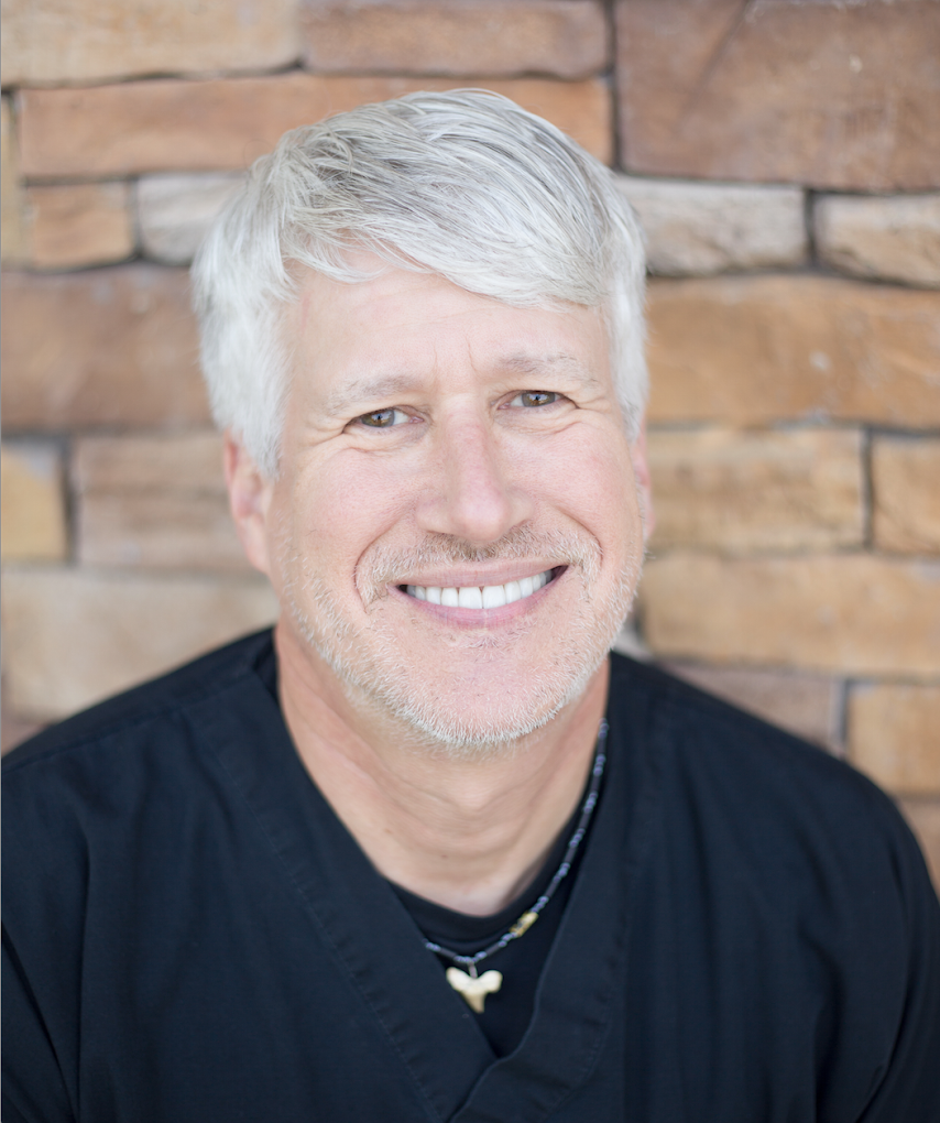 A man with gray hair and a beard is smiling in front of a brick wall. Your dentist near Lee's Summit, MO.