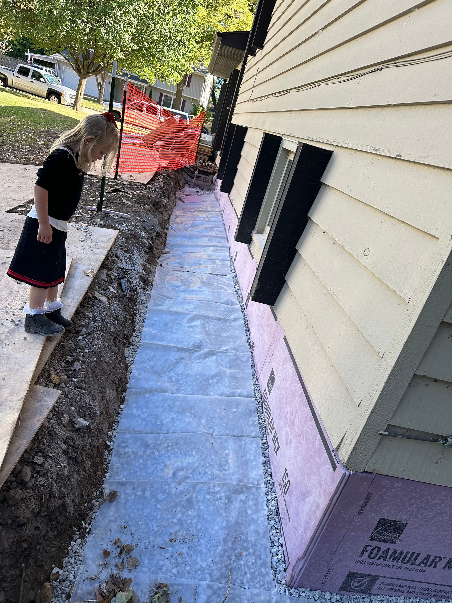 A little girl is standing in a trench next to a house.
