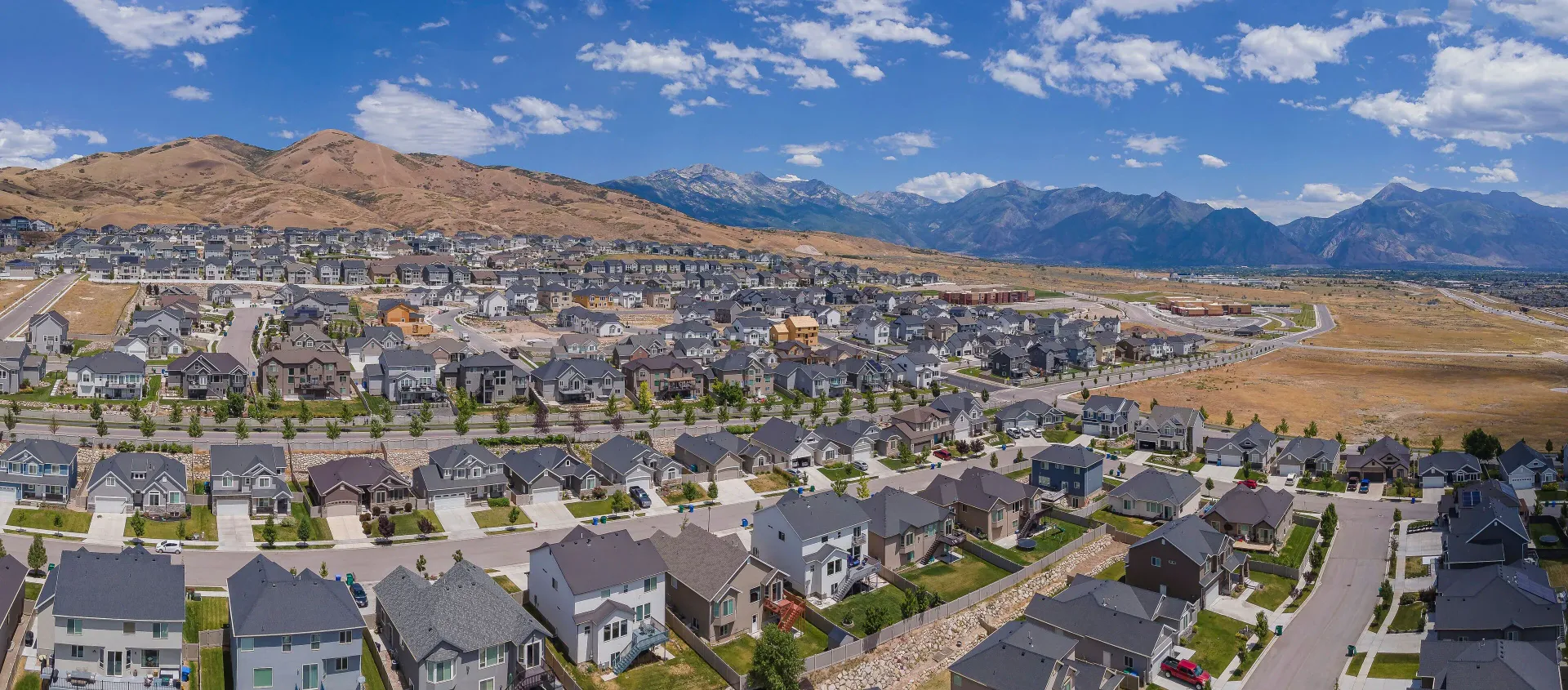 Panoramic view of a suburban neighborhood with gray houses, dry hills, and mountains under a blue sky