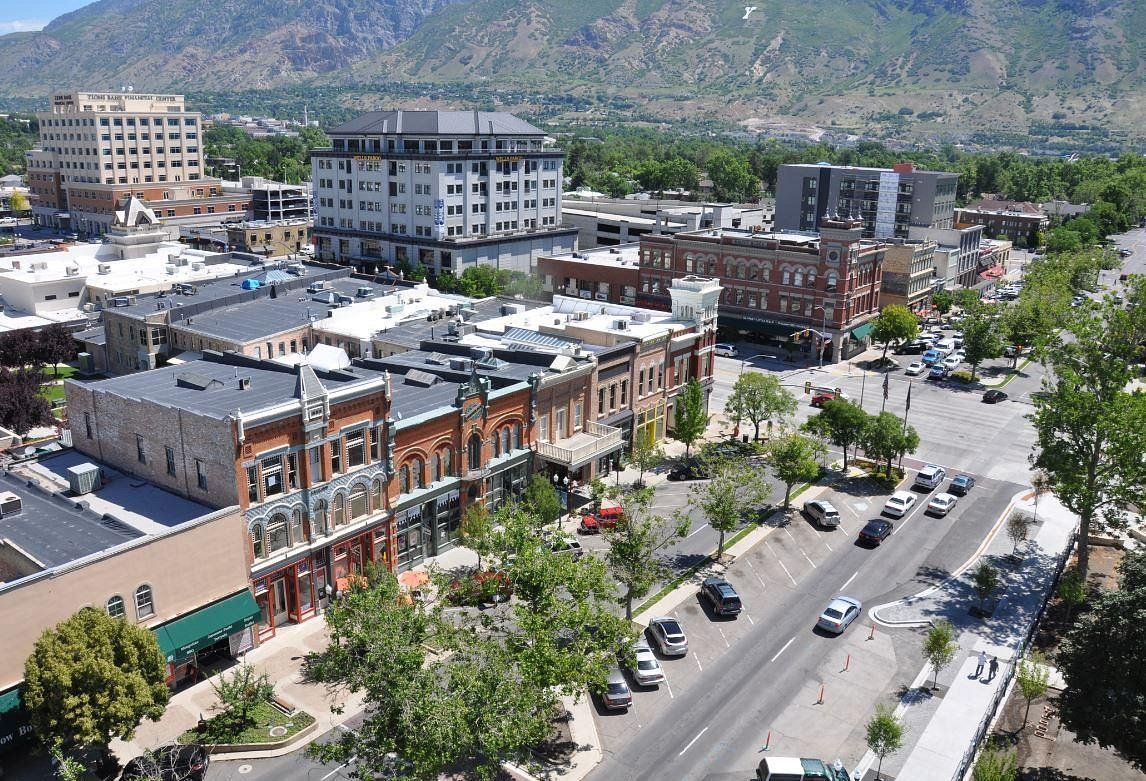 Downtown street with cars, trees, and low-rise buildings, backed by mountains under a clear sky