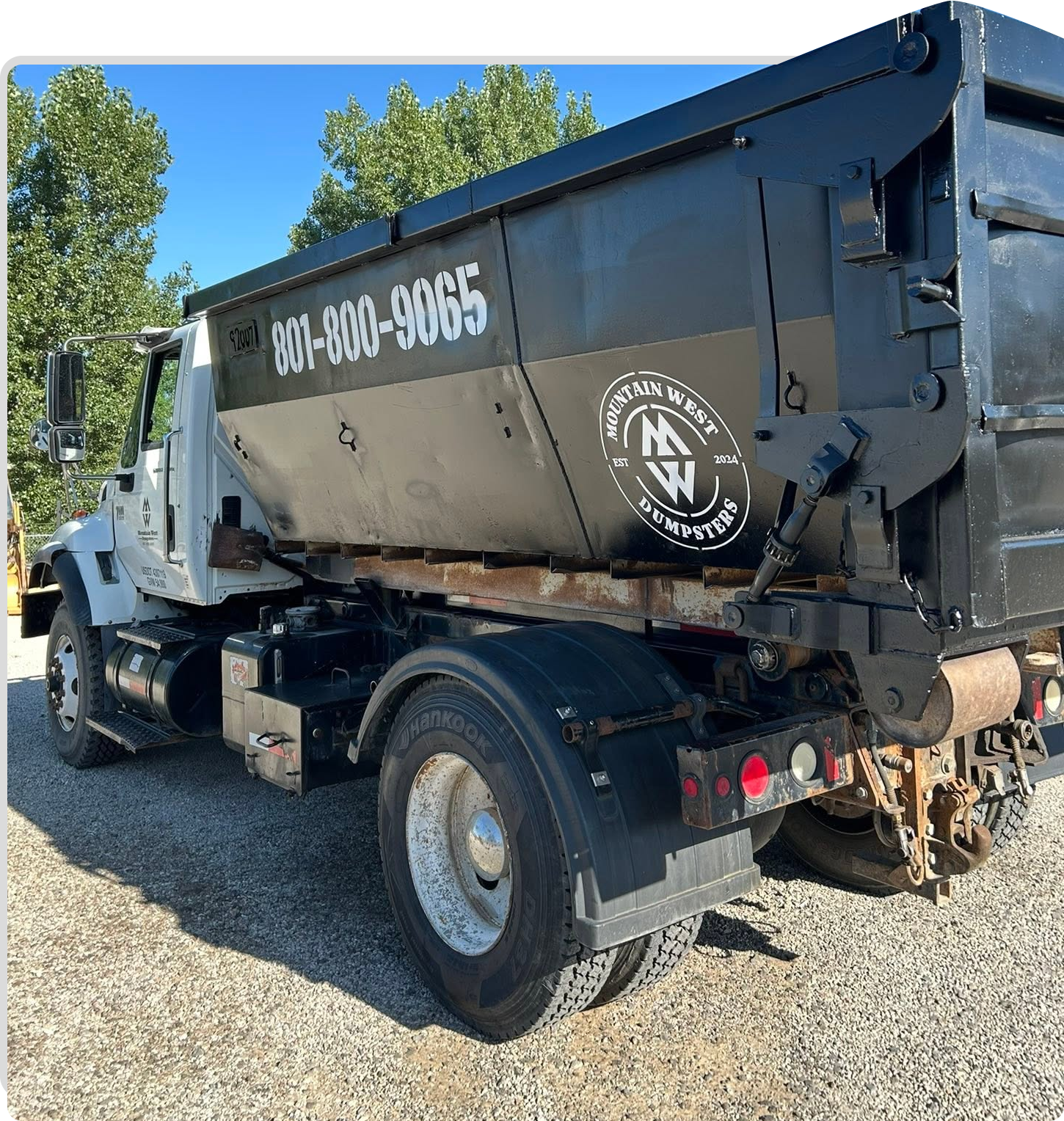 Black dump truck parked on gravel under a blue sky, side view with company phone number visible