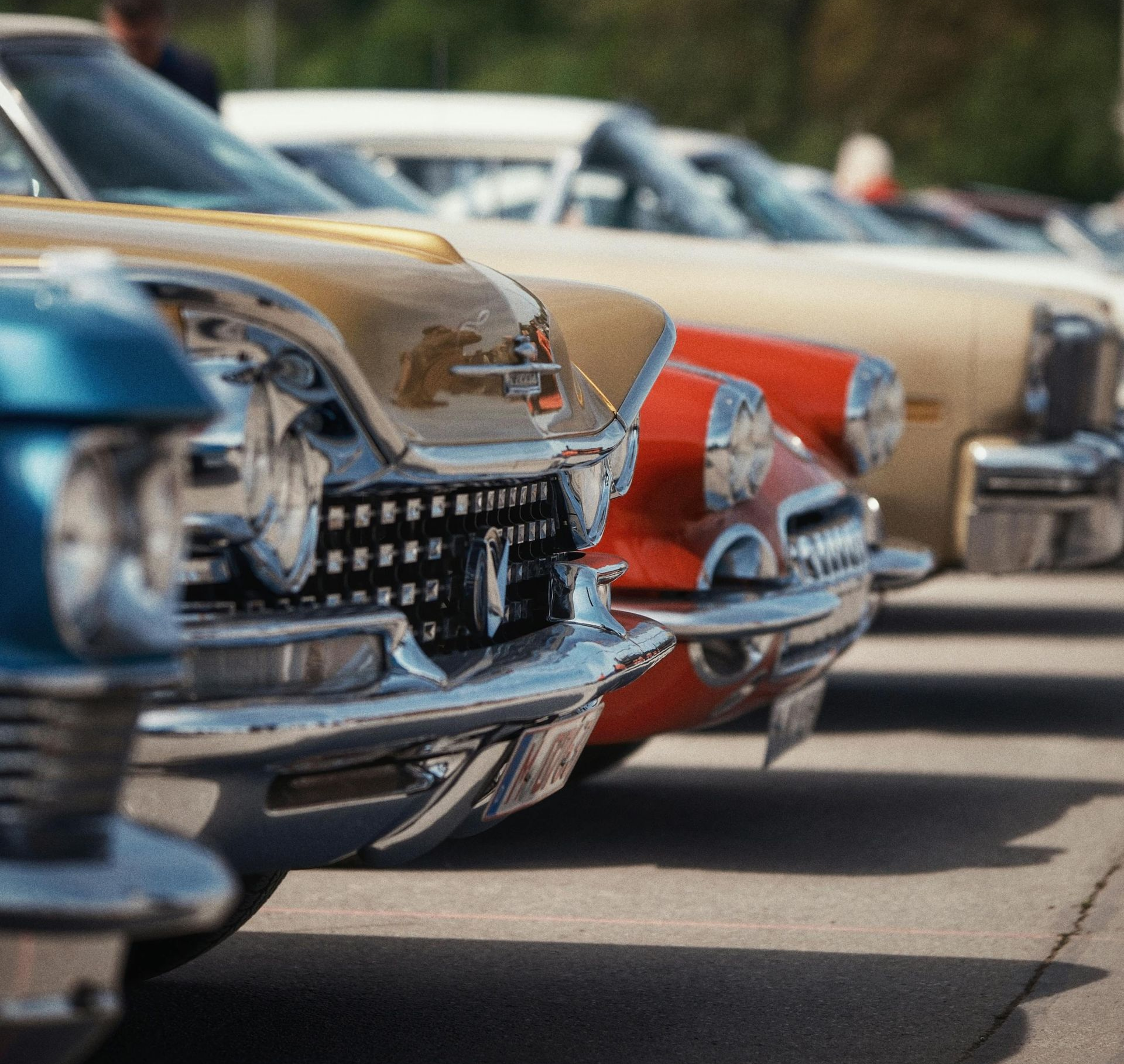 Row of classic cars parked close together, colorful fronts in a sunny outdoor lot.