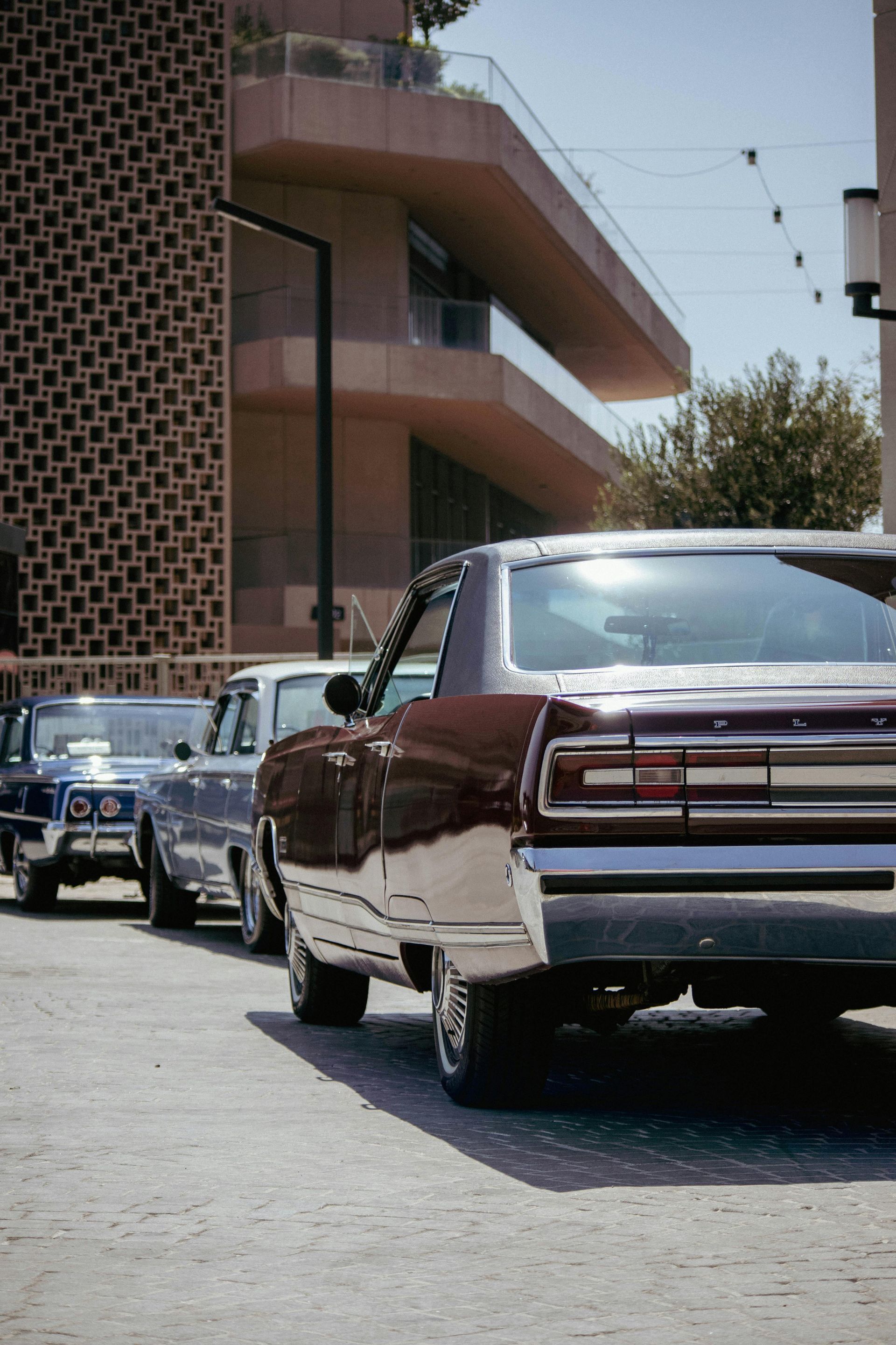 Classic cars lined up on a city street beside a modern building, rear view of a maroon convertible in front.