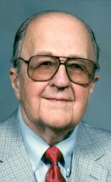 Portrait of a man in glasses, wearing a light blue suit and red tie against a plain blue background