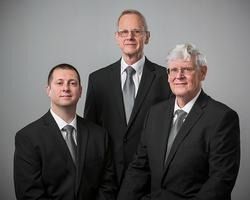 Three men in black suits pose against a gray studio backdrop.