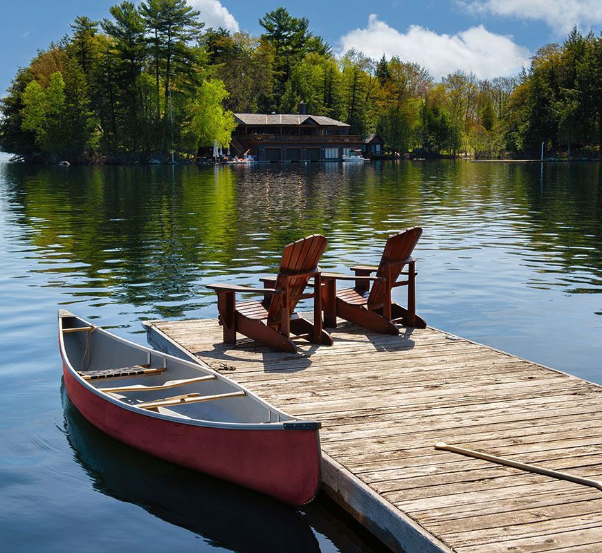 Wooden dock with two chairs beside a red canoe on a calm lake, with a wooded shoreline in the background