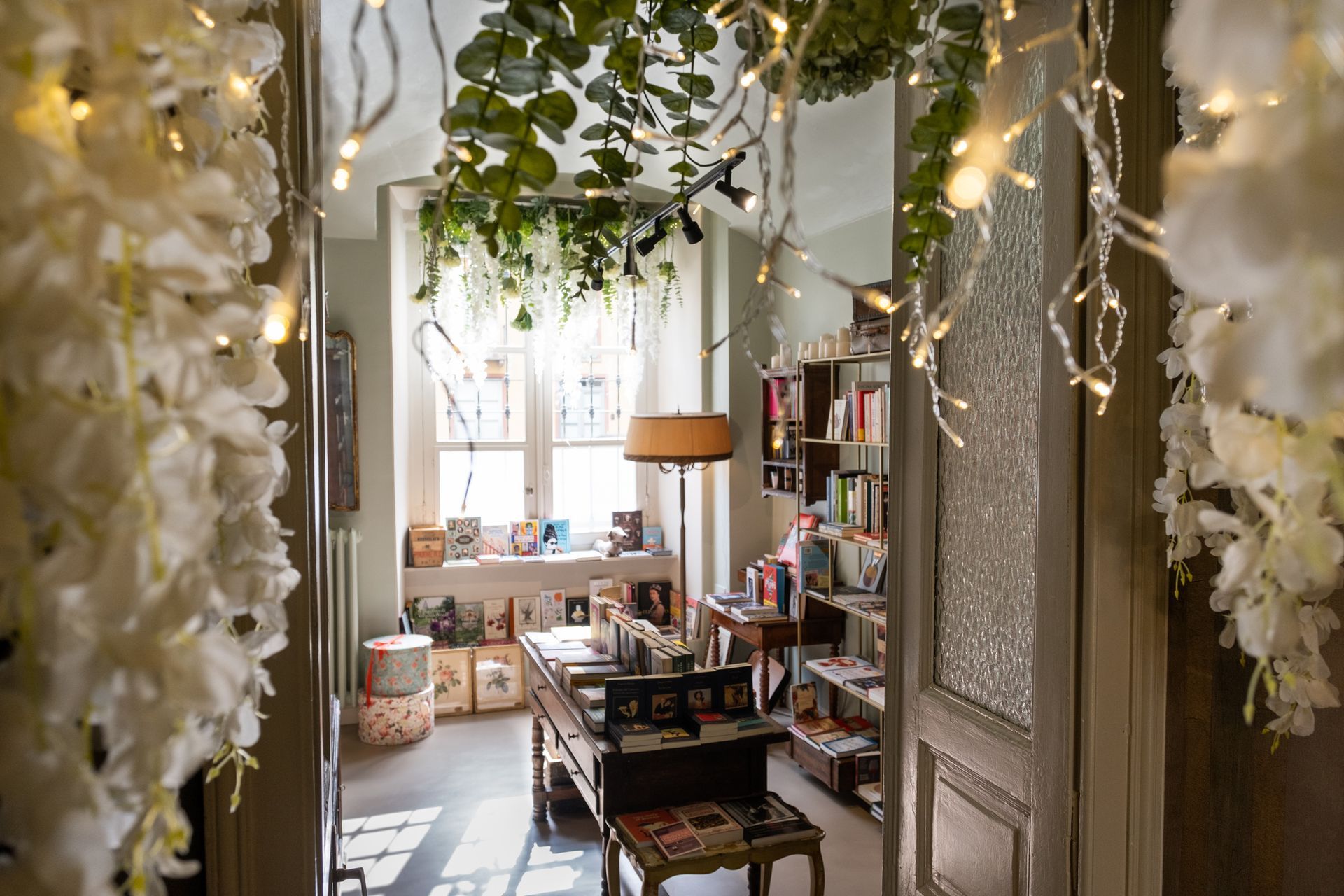 Accogliente corridoio di una boutique, adornato di fiori, con luci decorative, scaffali e una scrivania sullo sfondo.
