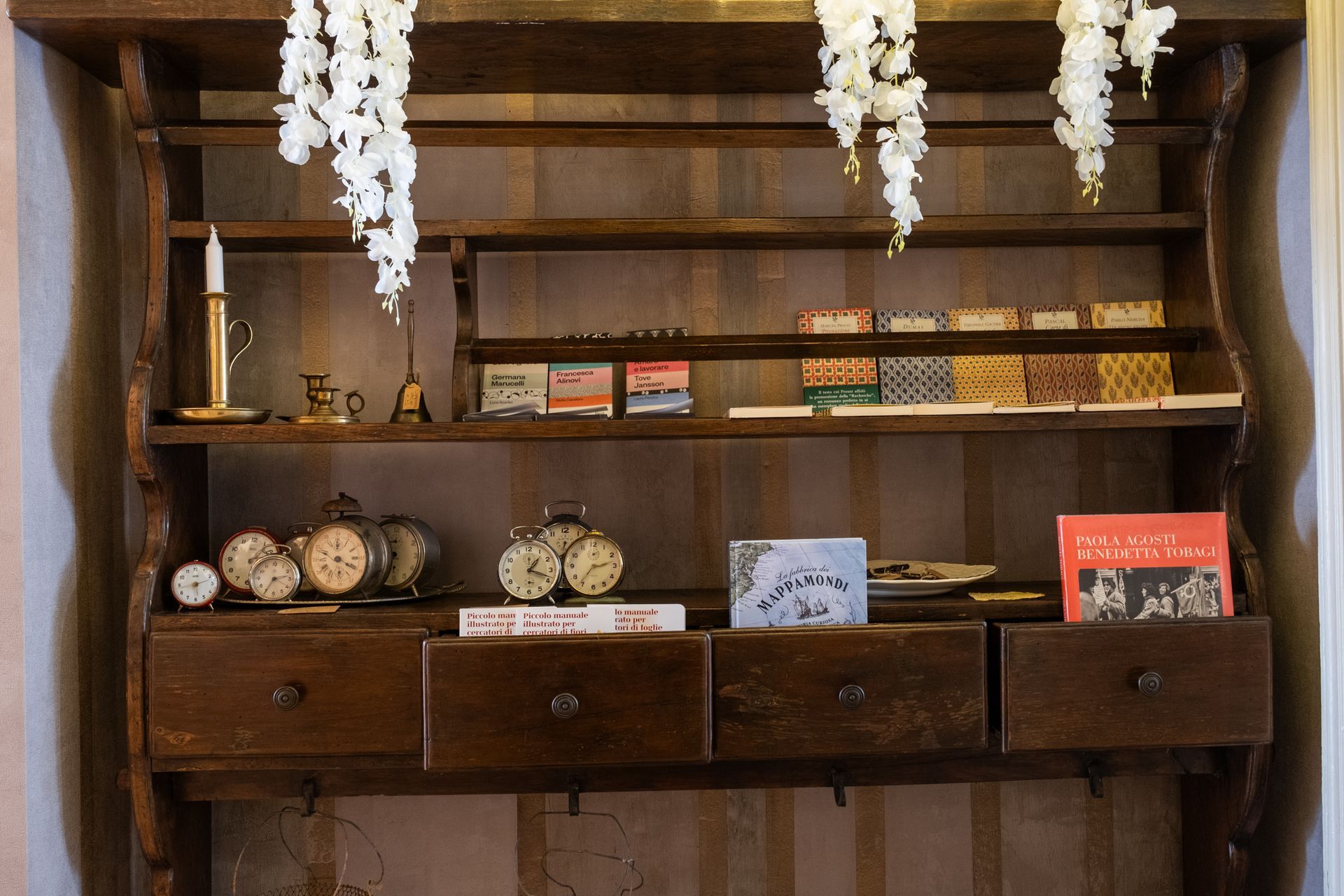 Scaffale in legno con libri, piccoli oggetti e fiori bianchi pendenti.