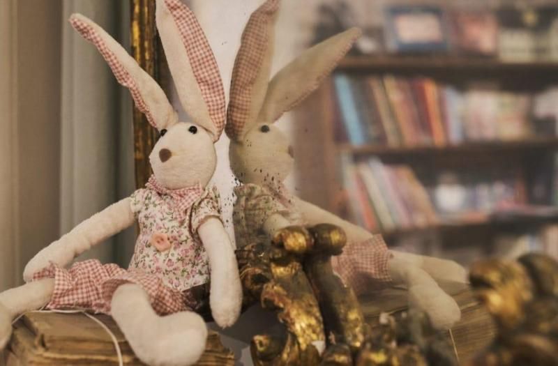 Due conigli di peluche fatti a mano, uno floreale e l'altro dorato, appoggiati su uno scaffale, con una libreria sfocata sullo sfondo.