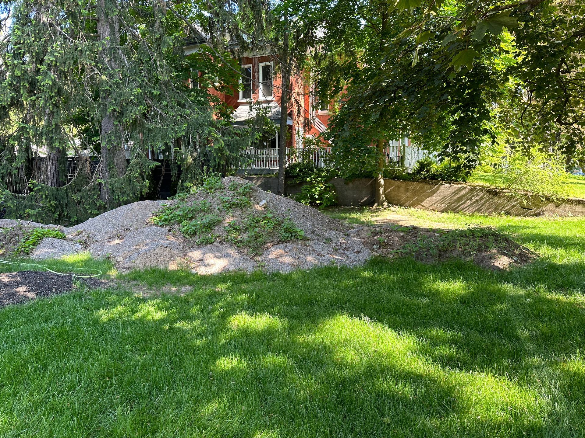 Tree-shaded yard with green grass, a gravel mound, and a house partially visible in the background