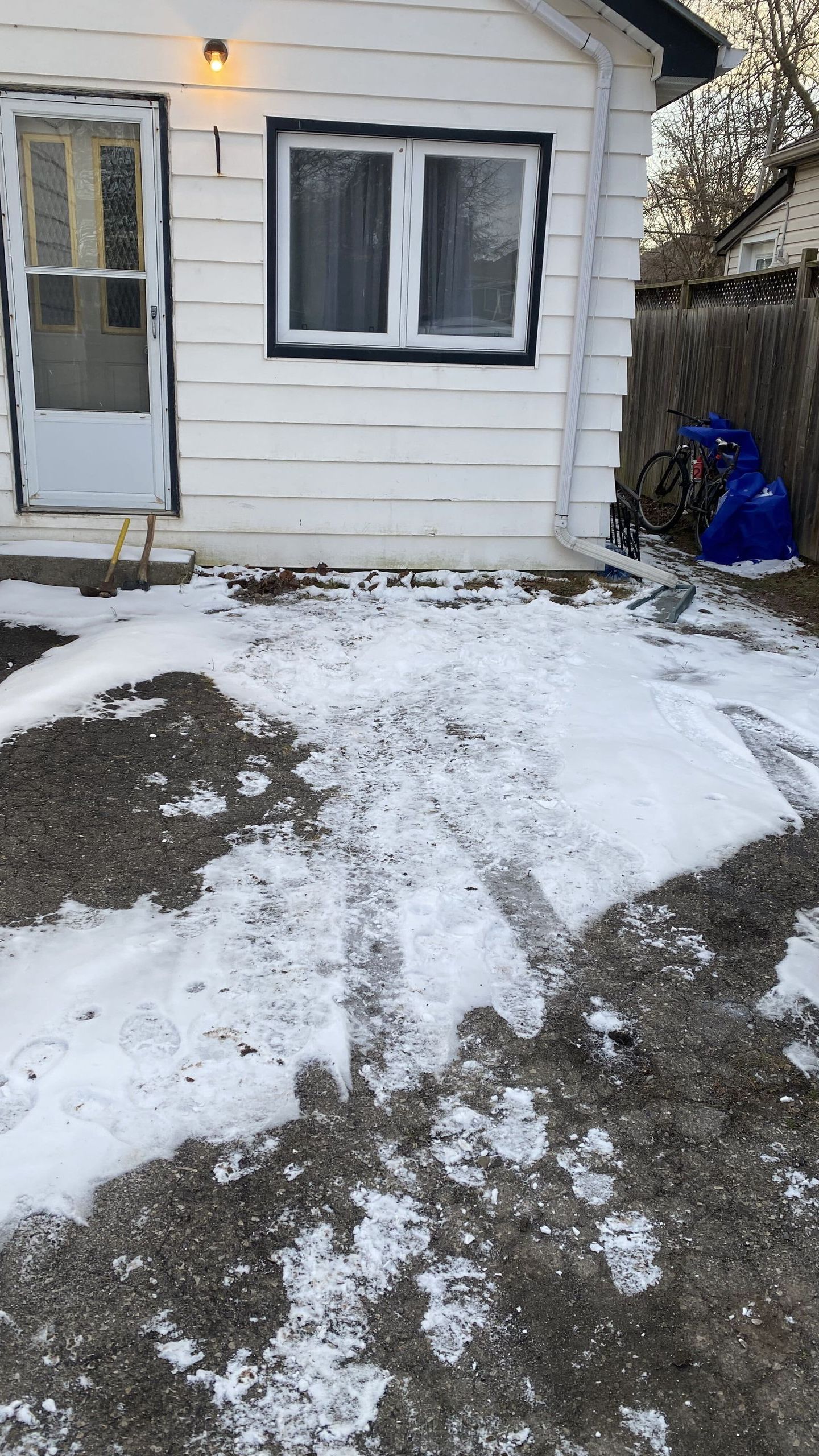 Snow-covered driveway beside a white house with a front door and window.