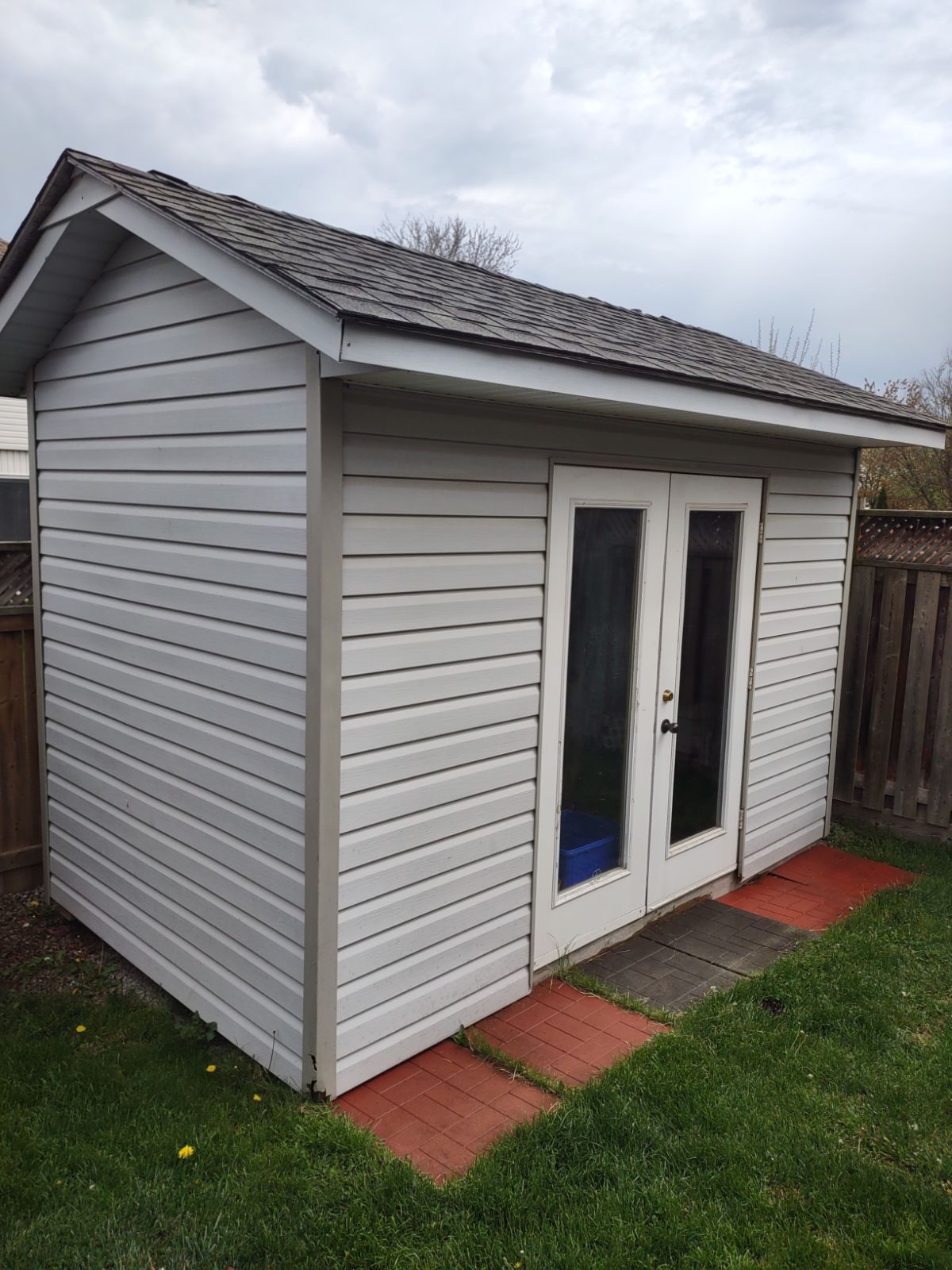 Small gray garden shed with white double doors and a tiled path in a grassy yard