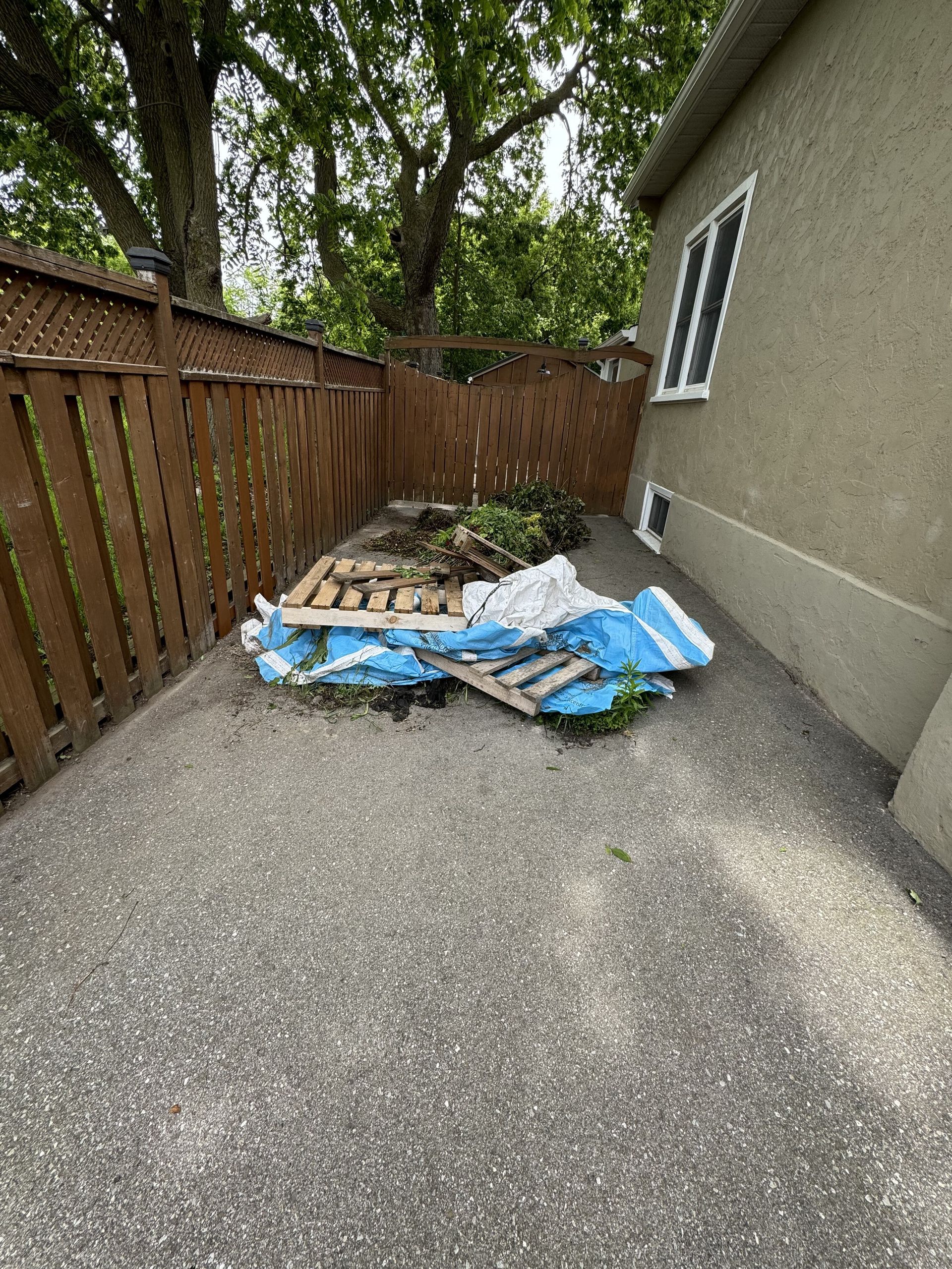 Backyard patio with a blue tarp and debris piled beside a house and wooden fence