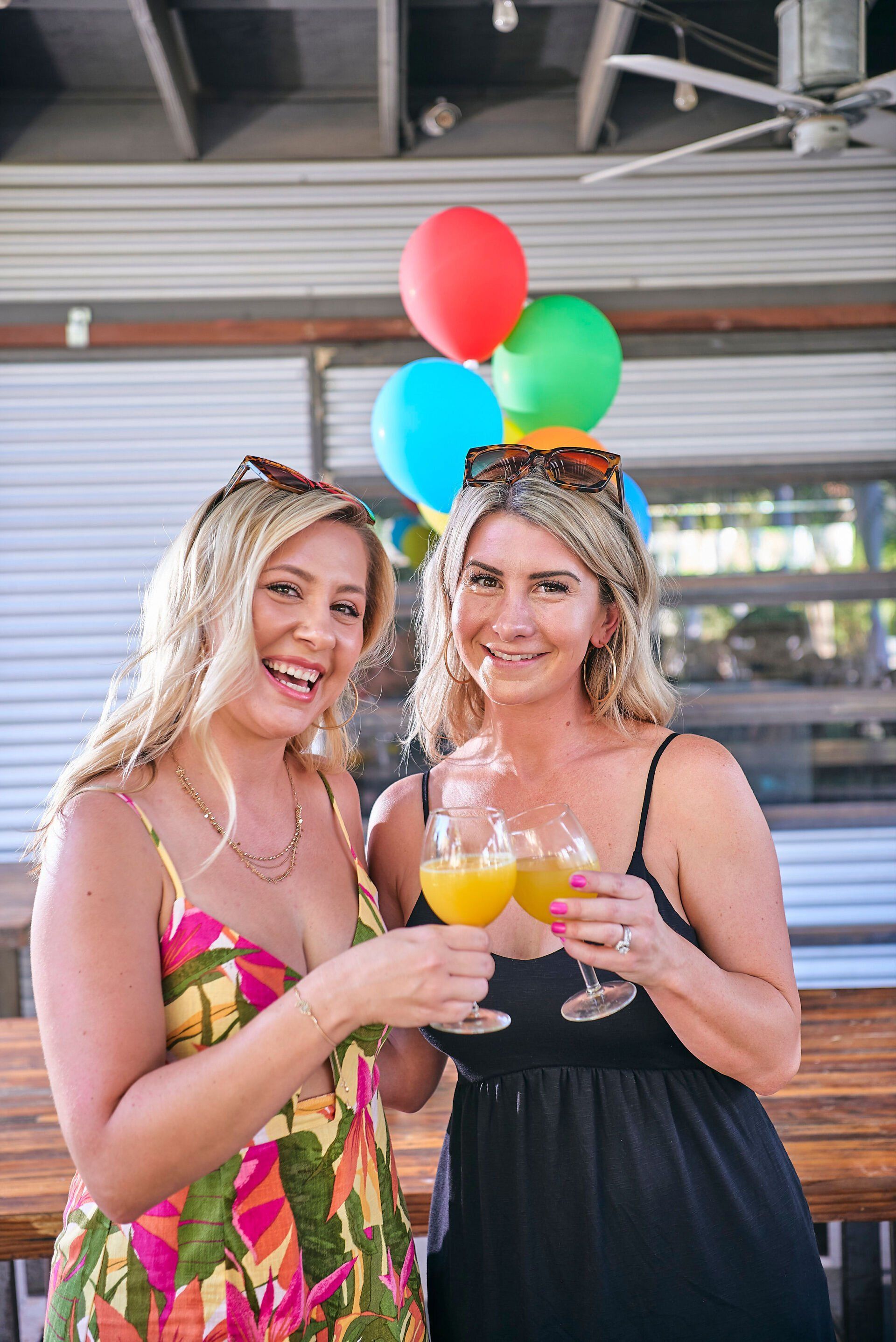 Two women are standing next to each other holding drinks and balloons.