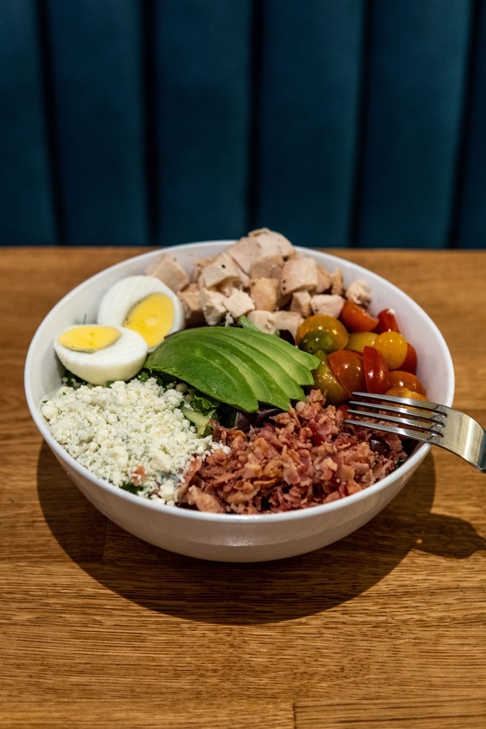 A bowl of food with eggs , avocado , bacon and tomatoes on a wooden table with a fork.