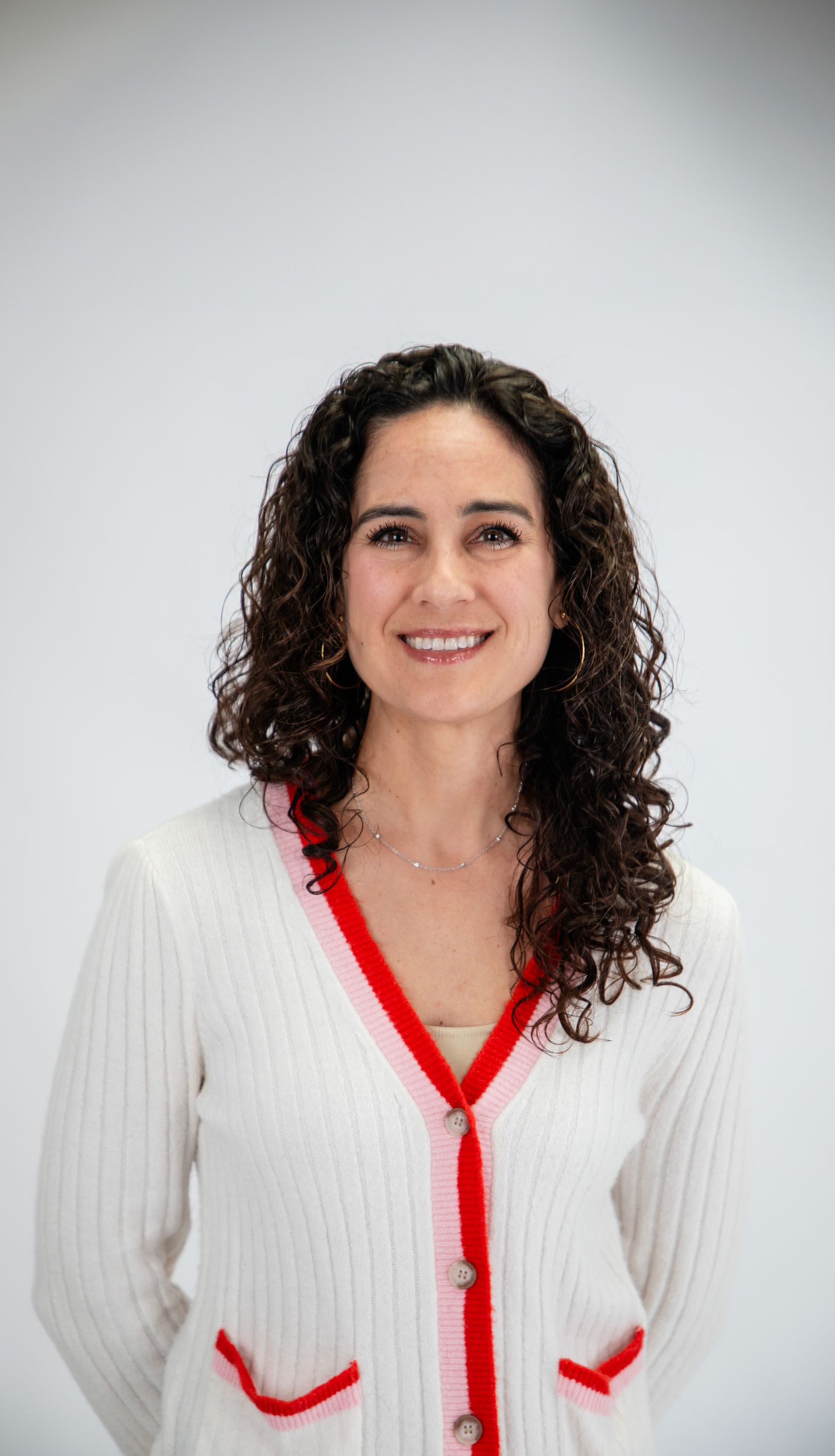 A woman with curly hair is wearing a white and red cardigan and smiling.