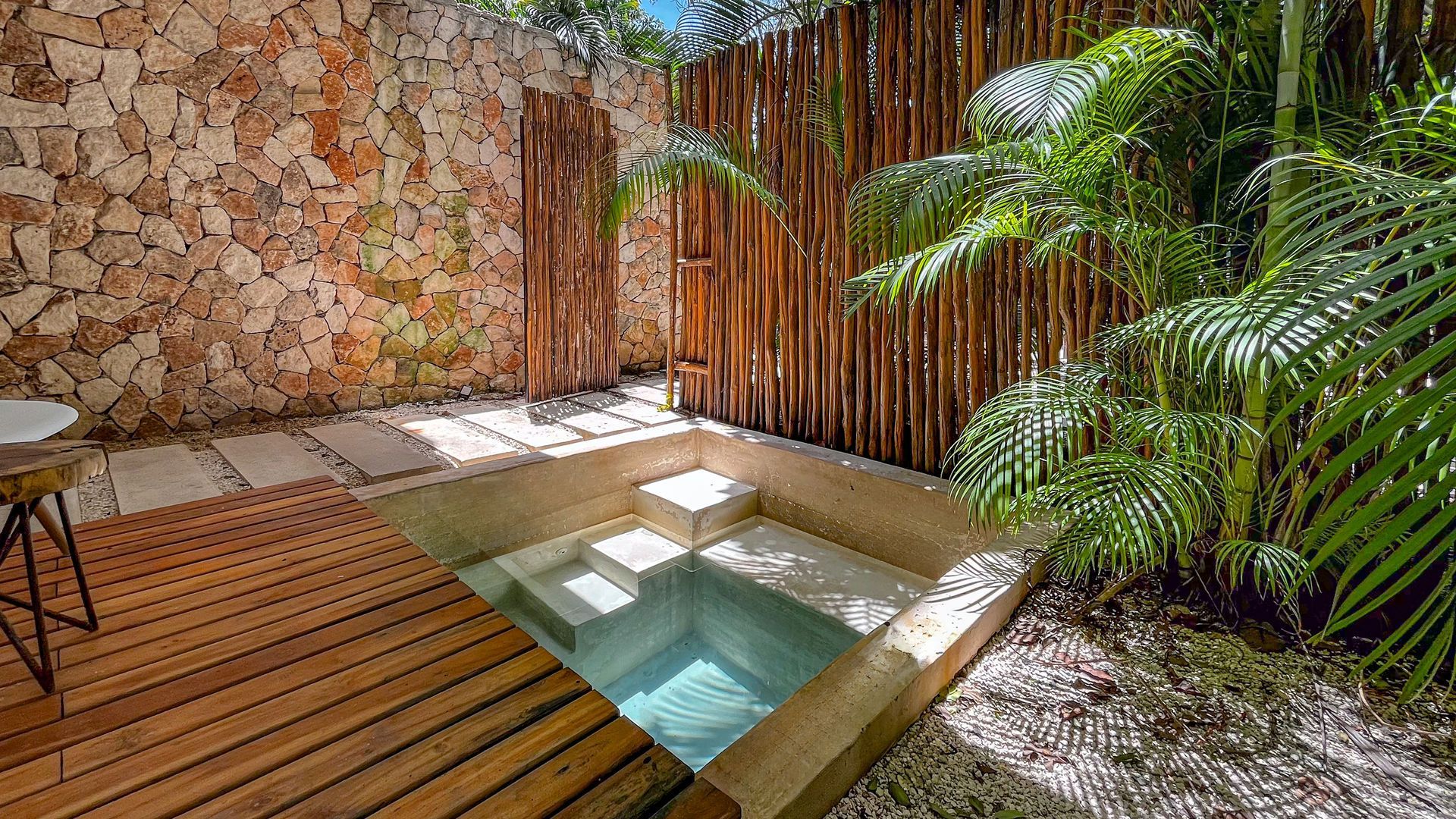 Jacuzzi al aire libre hundido rodeado de un muro de piedra, una valla de madera y una exuberante vegetación.
